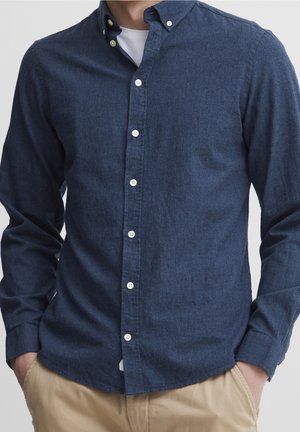 Homme portant une chemise bleu foncé à manches longues boutonnée sur un sous-vêtement blanc, les mains dans les poches d’un pantalon beige, torse visible sans visage.