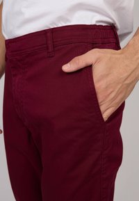 Homme portant un pantalon bordeaux et une chemise blanche, la main droite partiellement dans la poche avant, sur un fond neutre.