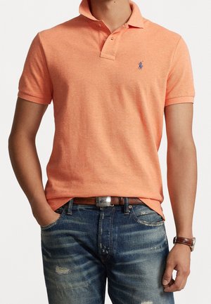 Polohemd aus weichem orangefarbenem Baumwollstoff, mit kurzen Ärmeln, zwei Knöpfen und gesticktem Logo. Getragen mit zerrissenen blauen Jeans.