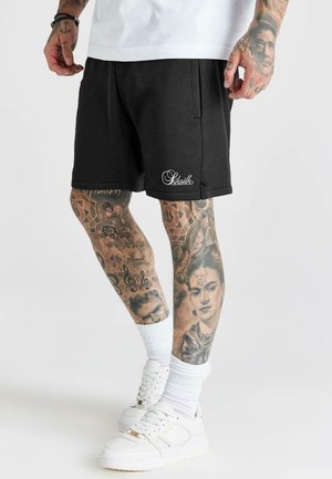 Mand iført sorte shorts med "Siksilk"-logo, hvide sokker og hvide sneakers, der viser kraftigt tatoverede ben og hænder.