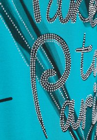 Turkoosi t-paita, jossa on musta graafinen kuvasto kimaltavilla strasseilla pyörivissä kuvioissa, tasaisen kankaan tekstuurin päällä.