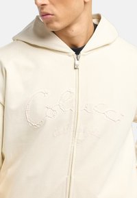 Creme zip-up hoodie med et broderet logo i et tekstureret design, der har en hætte og en sølv lynlås. Blødt, glat stof.