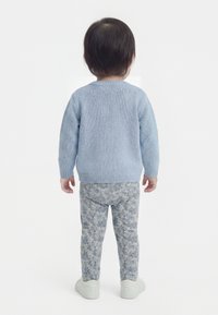 Kleinkind, von hinten, trägt einen hellblauen Pullover, gemusterte graue Hose und weiße Schuhe vor einem schlichten weißen Hintergrund.