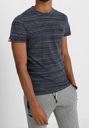 Man wearing dark blue striped short-sleeve T-shirt and light gray sweatpants with green drawstring, standing with one hand in pocket.