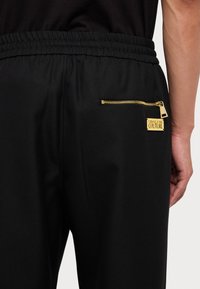 Black athletic pants featuring an elastic waistband, a zippered back pocket with gold hardware, and a label reading "Versace Jeans Couture."