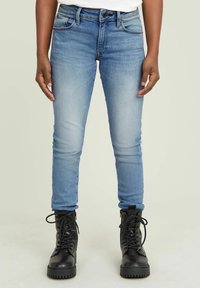 Person som bär ljusblå tajta jeans och svarta snörade kängor, stående mot en enkel beige bakgrund.