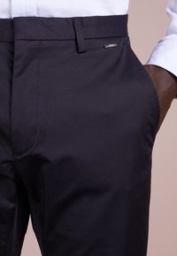 Navyblauwe broek van gladde stof, met een zijzak en een klein metalen logo-accent bij de taille.