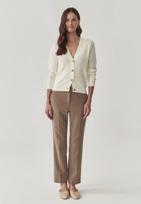 Weißer gerippter Cardigan mit Holzknöpfen, kombiniert mit braunen maßgeschneiderten Hosen und hellbeigen Slippern. Schlichtes, figuriertes Design.