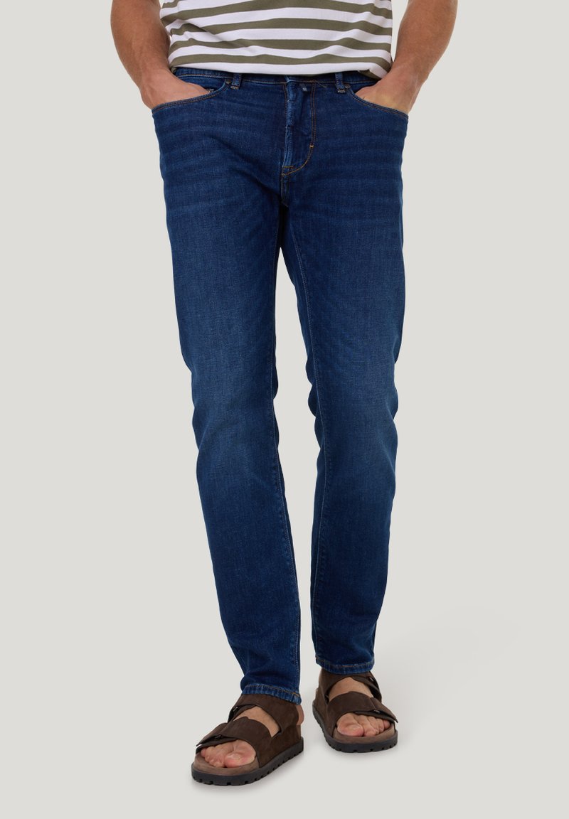 Donkerblauwe denim jeans met een rechte pasvorm, vijf zakken en contrasterende gele stitching, gecombineerd met bruine sandalen.