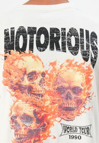 White graphic t-shirt featuring large black lettering "NOTORIOUS" above three colorful skulls with orange flames and a "WORLD TOUR 1990" label.