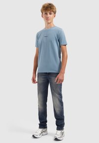 Tienerjongen staat, draagt een lichtblauw T-shirt, vervaagde blauwe jeans en witte sneakers tegen een effen lichte achtergrond.