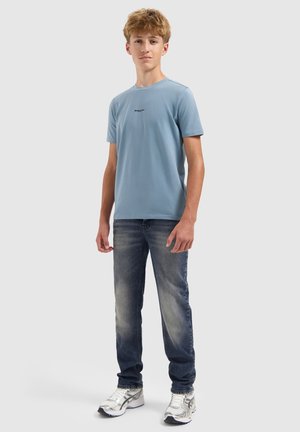 Tienerjongen staat, draagt een lichtblauw T-shirt, vervaagde blauwe jeans en witte sneakers tegen een effen lichte achtergrond.