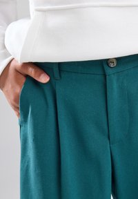 Türkisfarbene Hosen aus strukturiertem Stoff mit einem Bund und einem einzigen Knopfverschluss. Verfügt über Seitentaschen und Falten für eine maßgeschneiderte Optik.