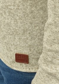 Pull en tricot beige avec un motif texturé, comportant une étiquette en cuir brun cousue à l'ourlet. Tissu en denim bleu visible en bas.