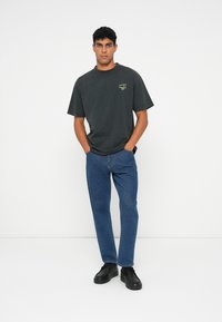 Donkergrijze katoenen T-shirt met korte mouwen, groene grafische afbeelding op de linkerborst, gecombineerd met blauwe spijkerbroeken en zwarte schoenen.