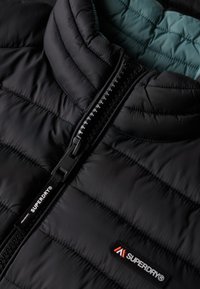 Zwarte gewatteerde jas met een doorgestikte textuur, een ritssluiting aan de voorkant met zwarte hardware, en een contrasterende teal binnenvoering. Voorzien van een logopatch.