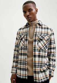 Sorte og hvide ternede flannelskjorte med brune accenter, der har to brystlommer og knaplukninger. Båret over en beige højhalset trøje.