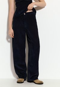 Svetlikaste široke hlače iz kordura v temno modri barvi z teksturirano rebrasto površino. Imajo sprednje zapiranje na gumb in žepov.