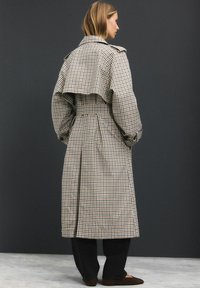 Beige und braun karierten Trenchcoat mit einem Rücken im Cape-Stil, weiten Ärmeln und einem taillierten Gürtel. Der Saum reicht bis zur Mitte der Wade.