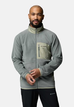 Mann mit Bart, der eine graue Fleecejacke mit Reißverschluss, hellbeigen Akzenten und Columbia-Logo trägt, steht mit verschränkten Händen.