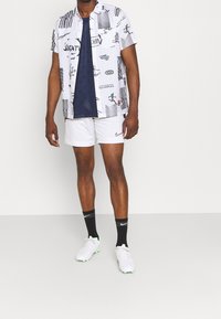 Bílá košile s krátkým rukávem s černými grafickými potisky, tmavě modrý síťovaný top pod ní, bílé šortky, černé ponožky a bílé sportovní boty se zelenými akcenty.