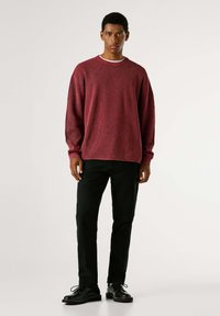 Roter, strukturierter gestrickter Pullover mit rundem Ausschnitt, kombiniert mit schwarzen, schmal geschnittenen Hosen und schwarzen Schnürschuhen.