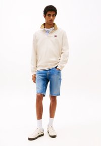 Kreemivärvi pullover, millel on kaelusega disain, koos helesiniste teksashüppesärkide ja valgete sportkingadega. Omab brodeeritud logo ja triibustikku.