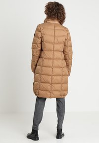 Abrigo acolchado largo beige con diseño acolchado, cuello alto y acento de logo negro en la manga. Combinado con pantalones grises con patrón y botas negras.