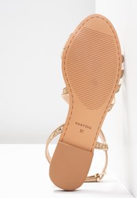 Semelle de sandale tournée vers le haut montrant une semelle en caoutchouc beige taille 37, des brides pailletées dorées et un petit talon sur un fond blanc en coin.