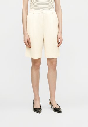 Persona vistiendo pantalones cortos hasta la rodilla de color crema, una camiseta sin mangas beige con rayas finas y tacones slingback negros de punta afilada.