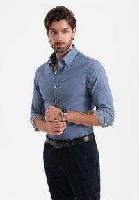 Camisa de botones azul claro con un acabado texturizado, mangas largas y puños plegables, combinada con pantalones de cuadros oscuros y un reloj plateado.
