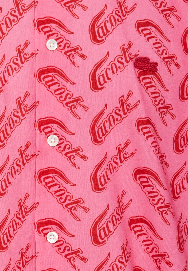 Roze stoffen shirt met een herhaald patroon van het rode Lacoste-logo. Het heeft witte knopen en een zachte textuur.