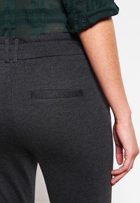 Grijze stretcht stoffen broek met een gladde textuur. Bevat een klein achterzakje met een horizontale zwarte accent. Close-up van de achterkant.