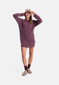 Nainen hymyilee, päällään vaaleanpunainen neulottu mekko, beige hattu, jossa on "R", ruskeat sukat ja beige tossut, poseeraa kädet hatun lähellä.