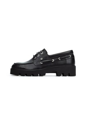 Zapato náutico de cuero negro con puntera redondeada, suela de goma negra gruesa, diseño con cordones y acentos de ojales plateados en los laterales.