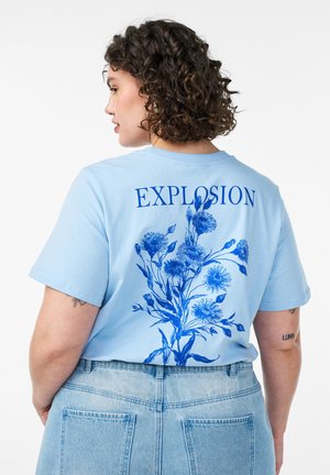 Person med krøllet hår iført en lyseblå t-shirt med blåt blomsterdesign og ordet "EXPLOSION" på bagsiden, kombineret med lyse denimjeans.