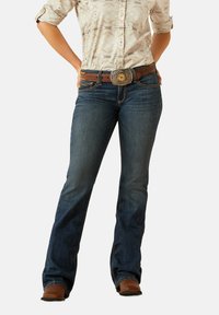 Dark blue denim jeans with a fitted waist and flared leg, accented by yellow stitching, worn with a patterned beige blouse and brown belt.