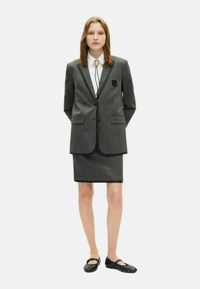 Grijze krijtstreep suit met een getailleerde blazer en een kokerrok. Bevat een zwart emblem op de blazer, gedragen over een witte blouse met een stropdas. Zwarte schoeisel.