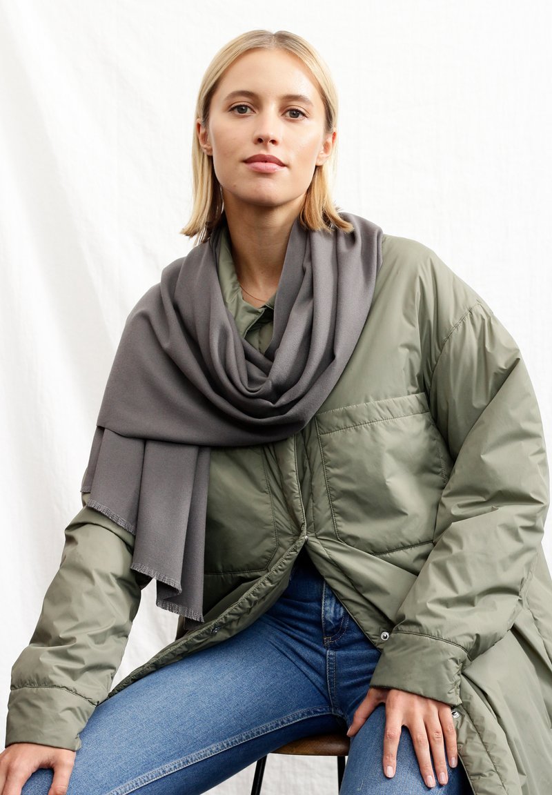 Écharpe grise drapée sur une veste en duvet verte, présentant une finition texturée et des bords effilochés. Le modèle porte un jean bleu, assis sur un tabouret.