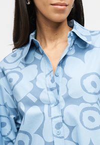 Γυναίκα που φοράει γαλάζιο πουκάμισο με μεγάλα αφηρημένα λουλούδια και ασημένια σκουλαρίκια κρίκους, κοντινό πλάνο στον λαιμό και το γιακά του πουκαμίσου.