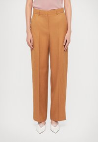 Pantalons larges en brown sur mesure avec une texture douce ressemblant à de la laine et une taille haute, associés à des chaussures blanches à bout pointu.