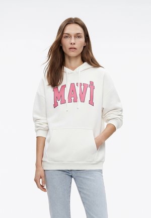 Frau mit langen braunen Haaren, die einen weißen Hoodie mit dem Schriftzug "MAVI" in großen pinken Buchstaben und hellblaue Jeans trägt, steht mit einer Hand in der Tasche.