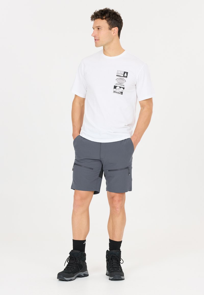 Mann steht mit den Händen in den Taschen, trägt ein weißes T-Shirt mit Grafik, graue Shorts mit Reißverschluss, schwarze Sportsocken und schwarze Wanderschuhe.