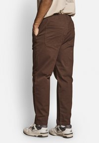 Bruine katoenen broeken met een relaxed fit, elastische tailleband, twee achterzakken en zijnaad. Gekoeld met grijze en zwarte sneakers.