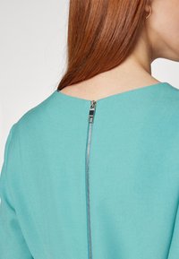 Femme aux longs cheveux roux portant un vêtement bleu sarcelle avec une fermeture éclair argentée visible courant le long de l'encolure arrière.