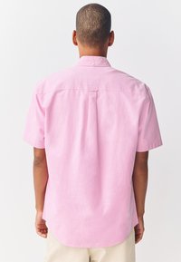 Camicia a maniche corte in tessuto rosa chiaro, con colletto button-down, dettaglio piega posteriore e orlo arrotondato.