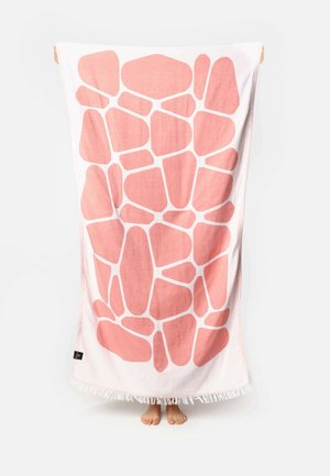 Personne tenant une grande serviette blanche avec un motif irrégulier rose rappelant la pierre, pieds visibles en bas, sur un fond blanc uni.