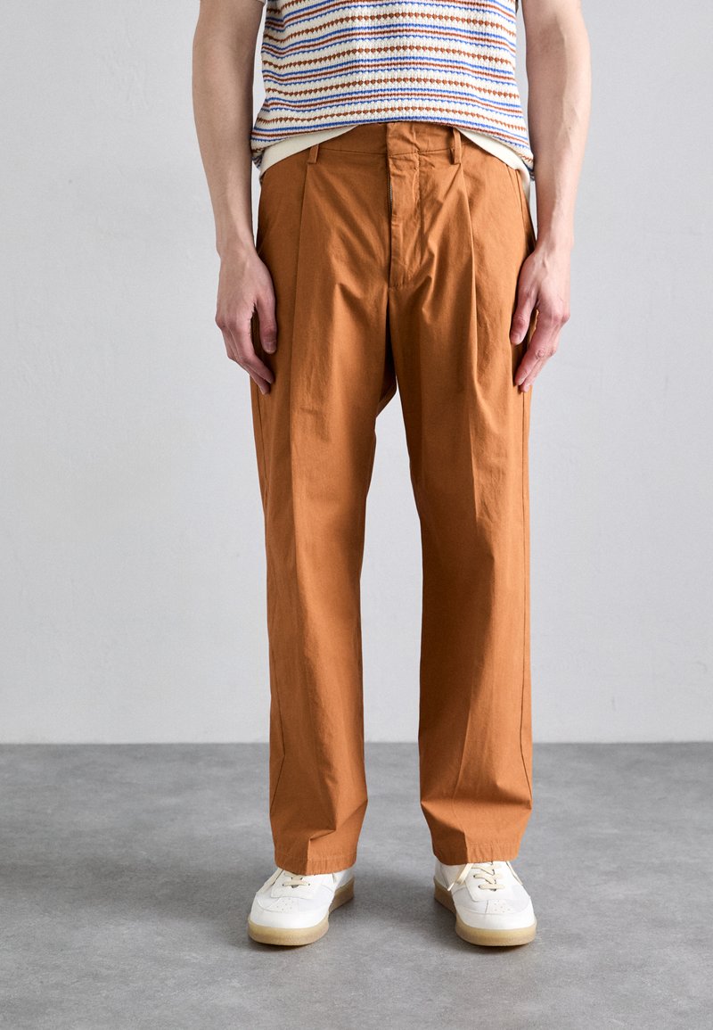 Pantalons plissés beige en coton léger, avec une coupe droite. Portés avec des baskets blanches sur un sol texturé gris.
