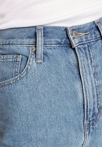 Os jeans em denim azul claro apresentam cinco bolsos, fechamento por botão e detalhes de costura visíveis ao longo das costuras. A textura é suave e ligeiramente desgastada.