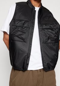 Svart nylonväst med dragkedjestängning fram, hög krage och flera fickor. Slät textur med ett subtilt mönster och en logotyp på bröstet.